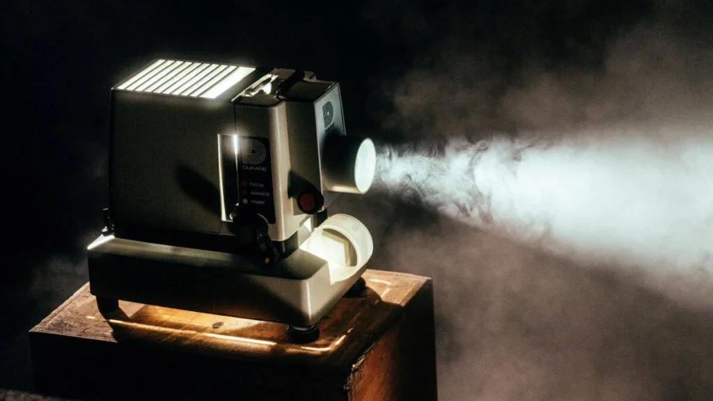 Alter Filmprojektor mit Metallgehäuse auf Holzunterlage, seitlich austretender Lichtstrahl mit sichtbarem Rauch in dunkler Umgebung