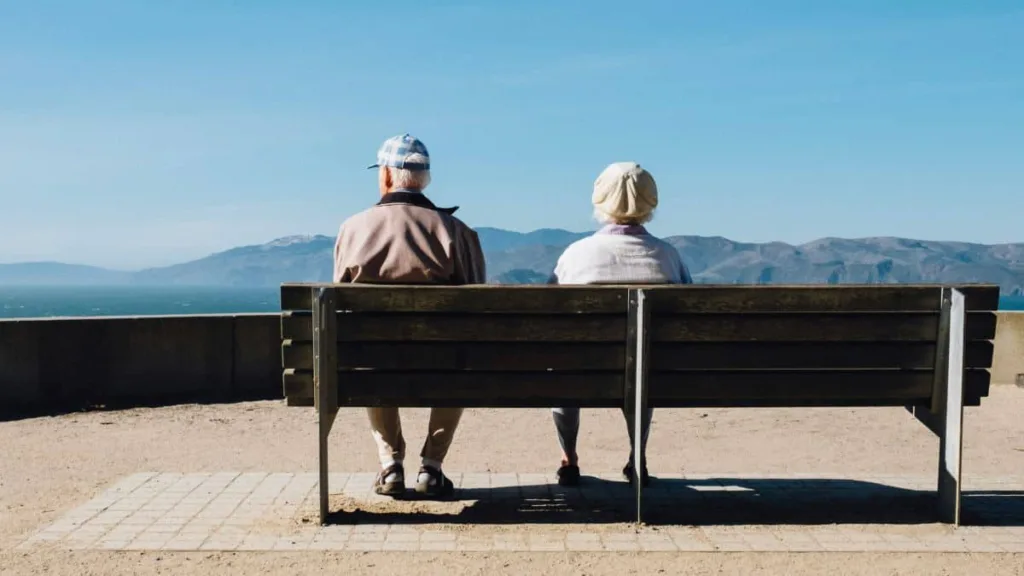 Älteres Paar sitzt nebeneinander auf einer Parkbank, von hinten fotografiert, und blickt gemeinsam über eine ruhige Landschaft mit weitem Horizont und freiem Himmel