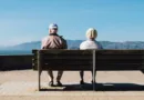 Älteres Paar sitzt nebeneinander auf einer Parkbank, von hinten fotografiert, und blickt gemeinsam über eine ruhige Landschaft mit weitem Horizont und freiem Himmel