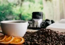 Weiße Kaffeetasse neben gerösteten Kaffeebohnen, Orangenscheiben und Kaffeekanne steht auf einem Tisch am Fenster