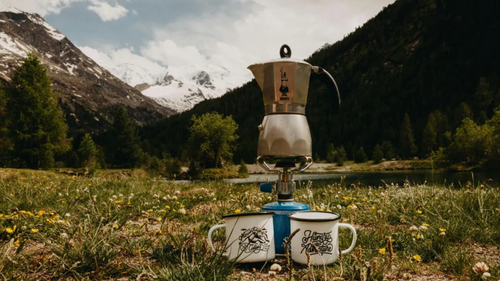 ein Kaaffeekocher steht auf einem Campingkocher und zwei Tassen stehen davor auf einer Wiese in den Bergen