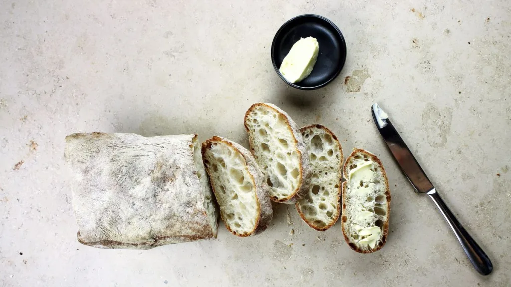 von einem rustikalen Brot sind 4 Scheiben abgeschnitten, daneben liegt ein Messer und eine schwarze Schale mit Butter