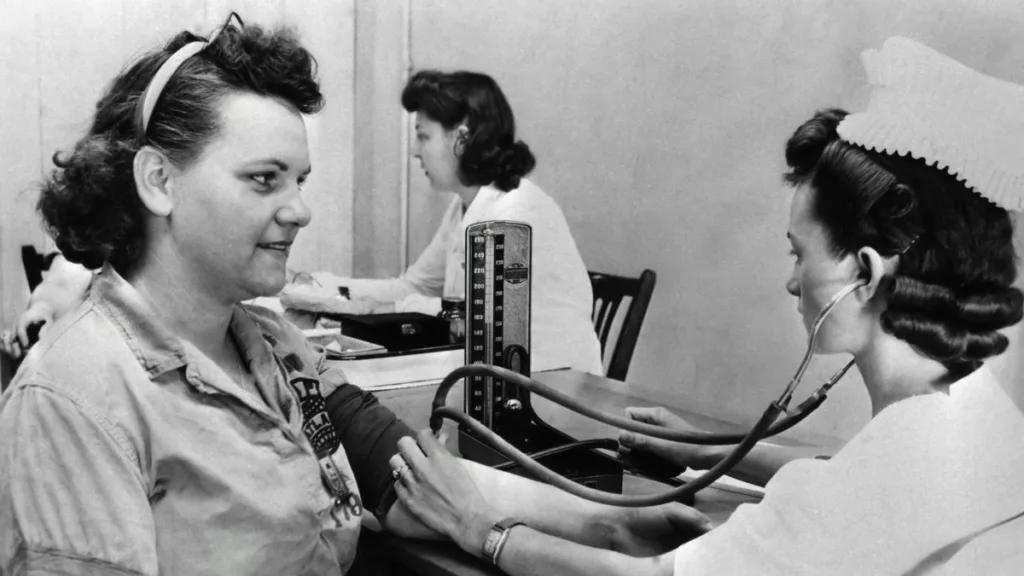 ein altes Schwarz-Weiß-Bild von einer Untersuchung, wo eine Krankenschwester den Blutdruck einer Frau misst