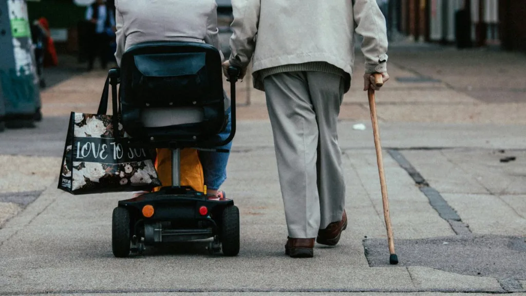 Ein älteres Pärchen gehen einen Weg entlang, sie im elektrischen Rohlstuhl und er zu Fuß mit Gehstock