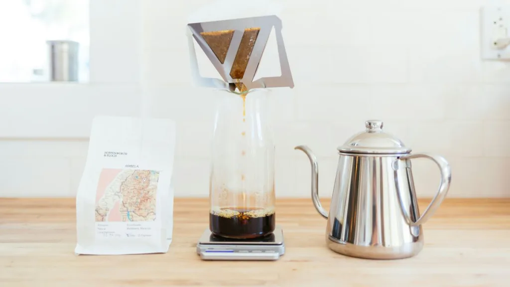 Kaffee läuft durch einen Filter in eine Glaskanne, daneben steht der Wasserkocher und die Kaffeetüte