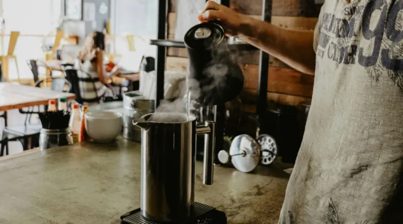 ein Mann gießt heisses Wasser aus einen Wasserkocher in ein Edelstahlgefäß