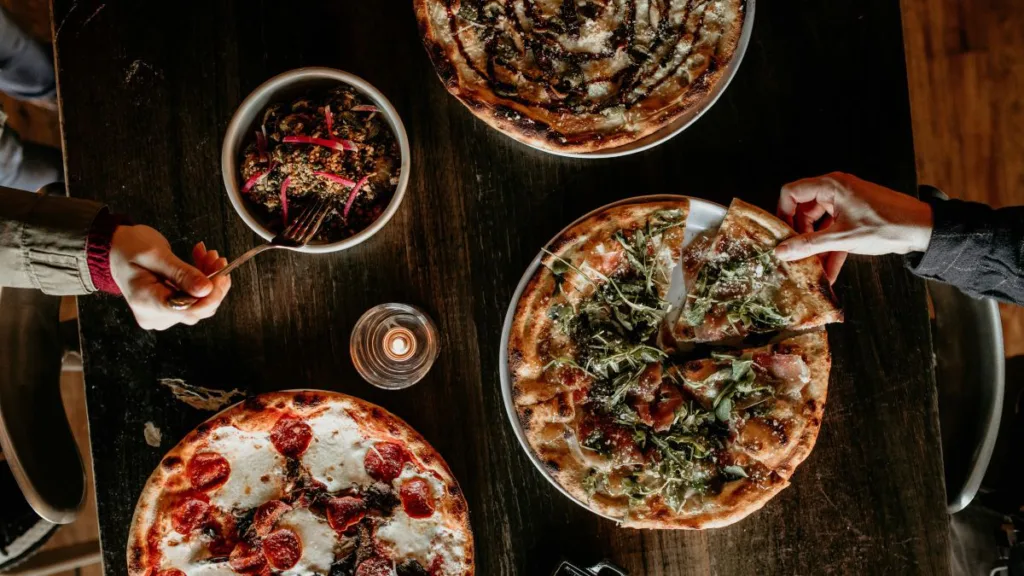 ein Essenstisch von oben, auf dem 3 Pizzen stehen und ein Salat, und eine Mnnerhand und eine Frauenhand greifen zu