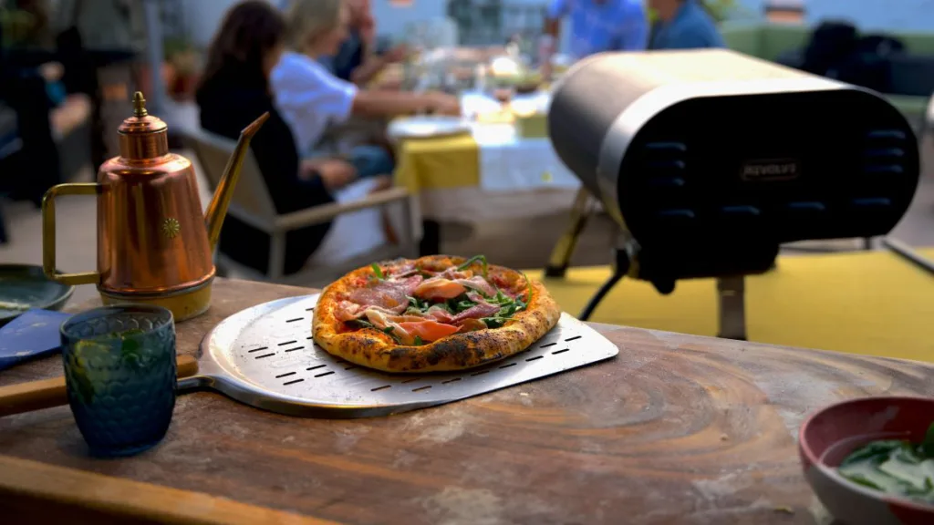 eine fertige Pizza aus einen Gas-Pizzaofen und im Hintergriund sitzen Menschen und Essen