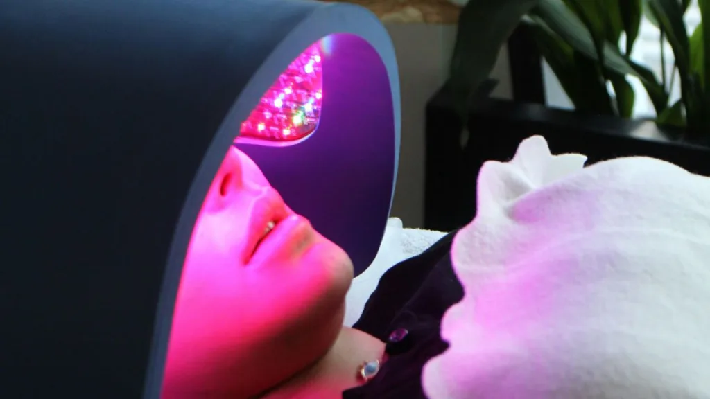 eine Frau liegt unter einem LED-Lichttherapiegerät, während das Gesicht mit pink-rotem Licht bestrahlt wird.